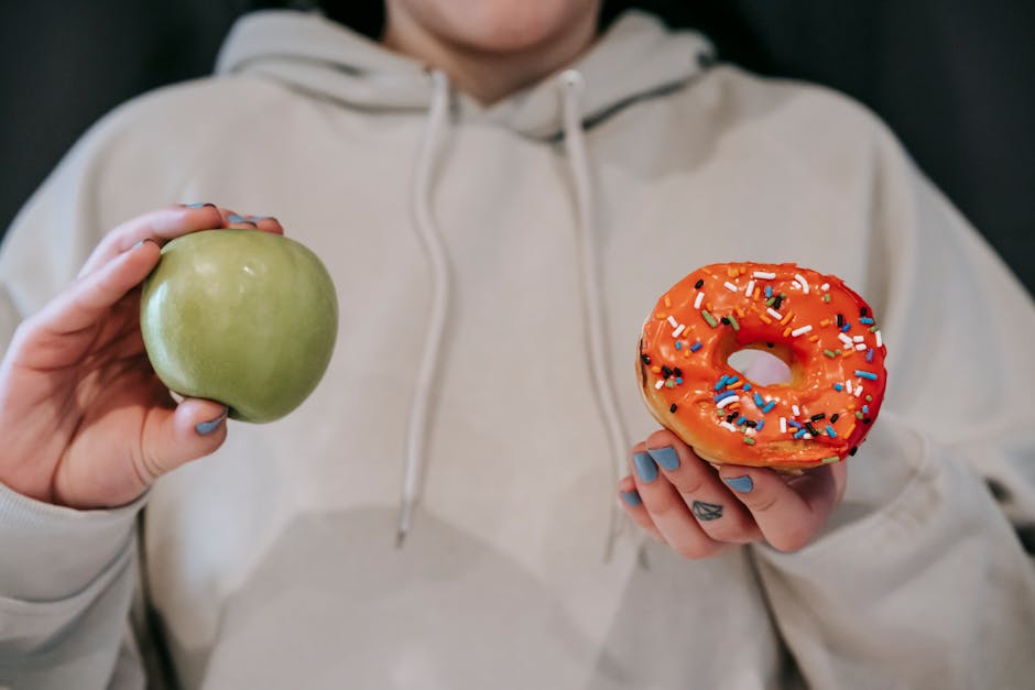 Crop unrecognizable female in loose hoodie showing healthy green apple and sweet delicious donut